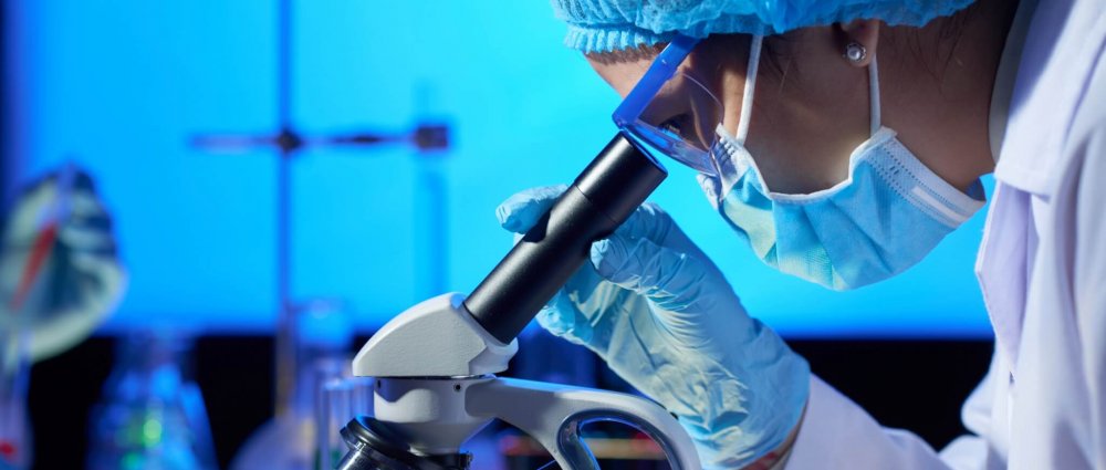 Profile view of concentrated Asian microbiologist examining sample with help of modern microscope while wrapped up in work at dim laboratory